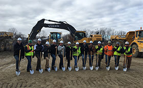 A group of safety workers in helmets hold shovels in front of some large equipment