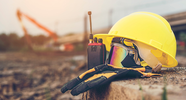 Construction hard hat, goggles, gloves, and walkie talkie displayed in the foreground of a construction site