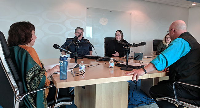 A group gathers around a table with microphones for a podcast discussion