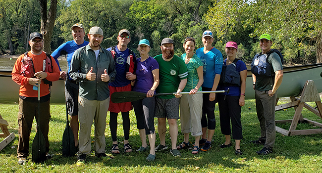 A group of outdoor enthusiasts gather in front of a canoe at a park