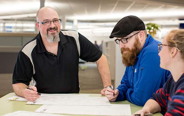 Profile photo of a man with glasses and a gray goatee smiling at a drafting table with two other colleagues