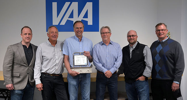 A group of six men gather in front of the VAA logo, one of them holds an anniversary award