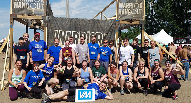 A group of employees gather outside in front of a Warrior Hangout Sign dressed in activity gear